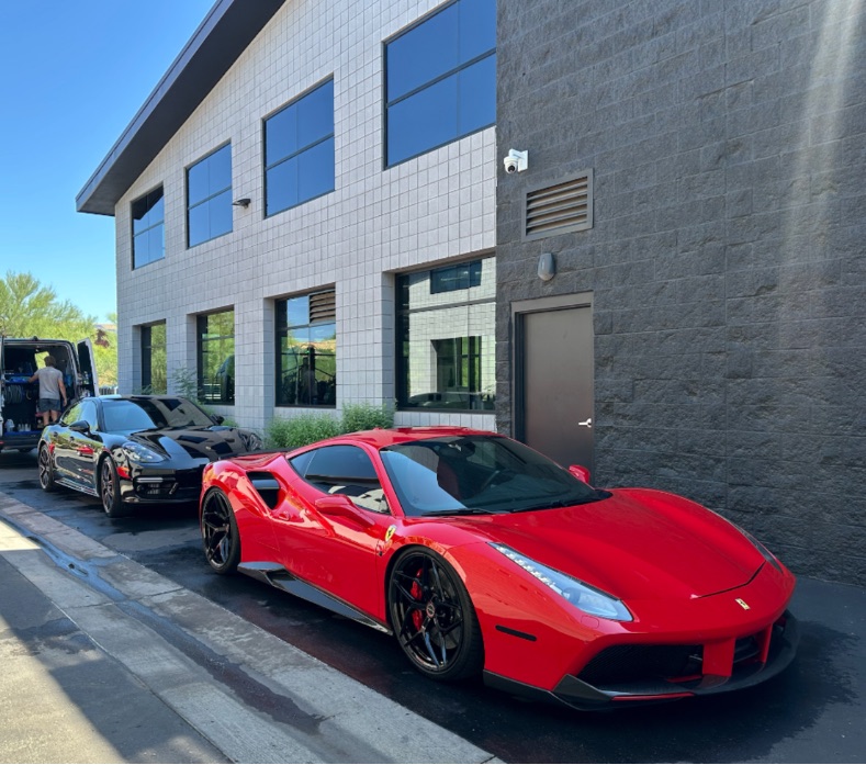The crew's van open mid-job on a commercial lot, a black Porsche Panamera and a red Ferrari parked beside it